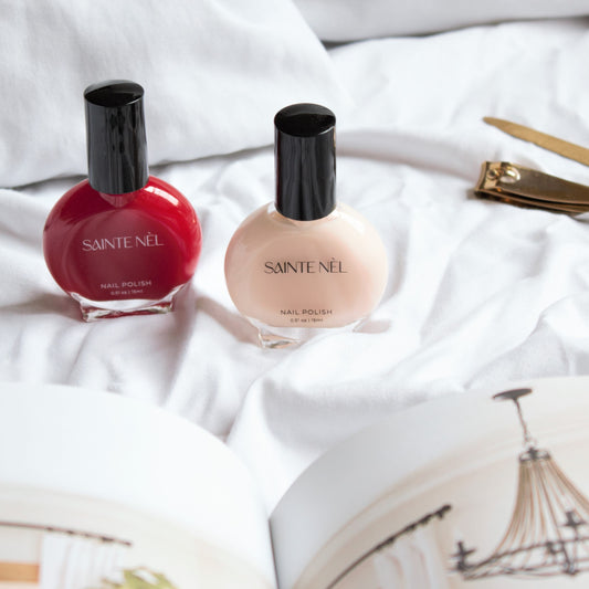 two bottles of the best vegan nail polish. one is red nail polish and the other is nude nail polish in a vintage nail polish bottle with a black cap. the front of the bottle says Sainte Nel. The back of the bottle says meticulously crafted non toxic vegan formulas created to deliver beautiful and healthy nails.