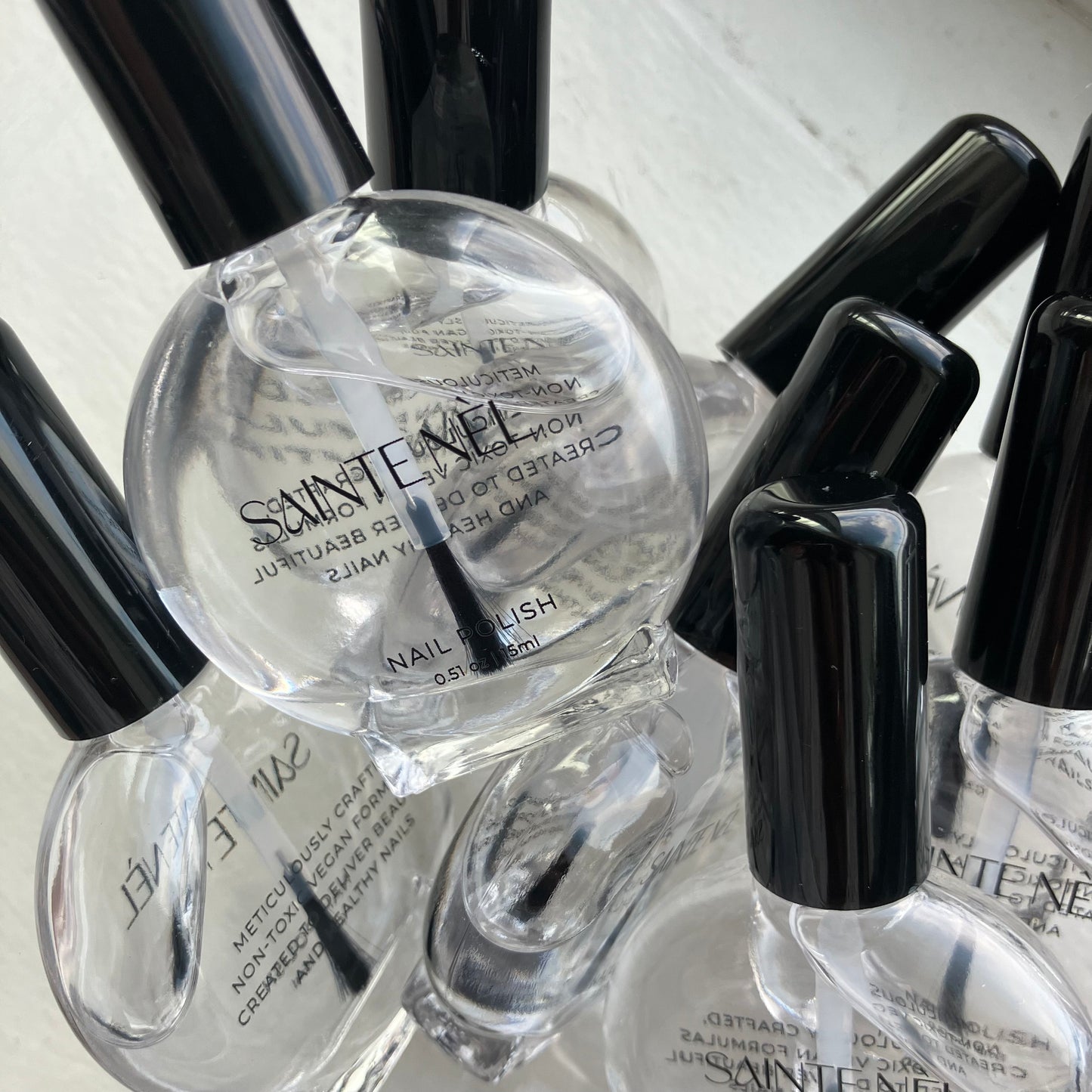 multiple bottles of a clear vegan nail polish in a vintage nail polish bottle with a black cap. the front of the bottle says Sainte Nel. The back of the bottle says meticulously crafted non toxic vegan formulas created to deliver beautiful and healthy nails.